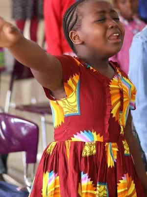 A young girl of African descent is captured mid-worship, with her eyes closed and arms raised in a gesture of praise. She wears a vibrant, patterned dress in shades of red, yellow, and orange. Behind her, blurred figures suggest a gathering. The lighting is soft with a slight overexposure.