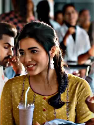 A woman with dark hair in a braid smiles and looks towards the viewer. She is seated in a busy restaurant, holding a drink in a plastic cup. The woman wears a yellow shirt with red accents. The background is blurred, with other people visible.