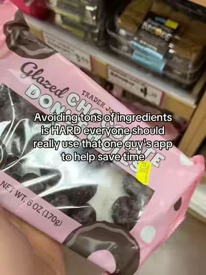 The image shows a close-up of a bag of Trader Joe's glazed chocolate donuts. Black glazed donuts fill the clear part of the bag. Text overlays the bag, mentioning avoiding ingredients and using an app.