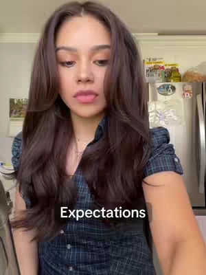 A woman with long, wavy brown hair is centered in the frame. She is looking downward with and wears a dark plaid shirt. The background includes a kitchen.
