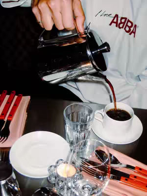 The image shows a close-up of a table setting with a person pouring coffee from a silver pot into a white coffee cup. The table is set with a white plate, a glass of water, and silverware. The person is wearing a white shirt with the words 'I Love ABBA' printed on it.