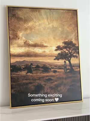 A framed canvas art piece, slightly angled, displayed on a white surface. The artwork depicts a landscape under a dramatic, glowing sky with a golden frame. In the foreground, the silhouette of a figure appears to be walking. In the distance there is a small tree with two figures. At the bottom is overlay text. The overall composition is warm-toned and evokes a sense of tranquility or anticipation.