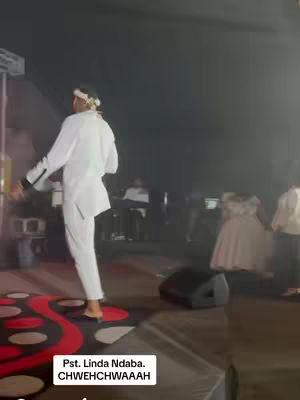 A person wearing a white suit and a headpiece is performing on stage. The floor has a red carpet design. On the bottom of the frame, there is text overlaid.