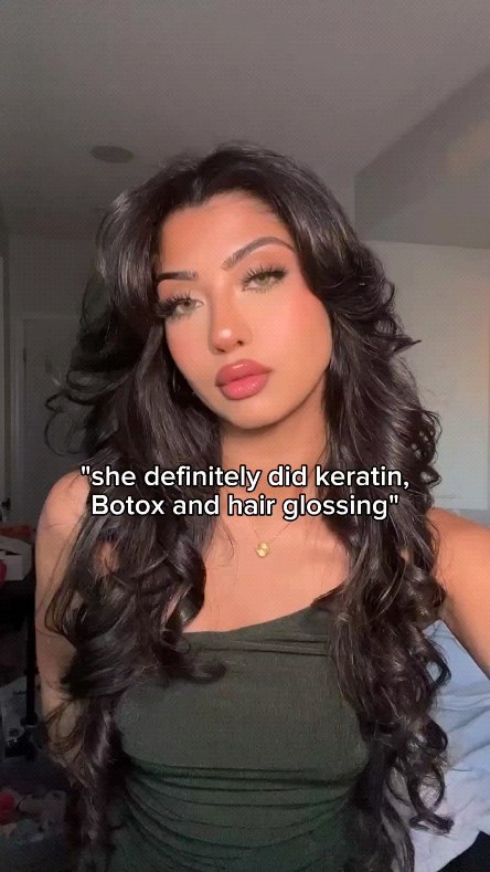 A young woman with long wavy black hair is centered in the frame, with a medium skin tone, full lips, prominent cheekbones. She is wearing a dark green top and a gold necklace. The background is blurred, with a soft lighting. A text overlay is present on the image.