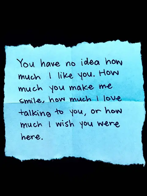 A close-up shot of a light blue, rectangular piece of paper with ripped edges, centered against a black background. The paper has handwritten text in black ink. The text is a personal message expressing feelings of affection and longing. The overall composition is simple and intimate, with a focus on the handwritten note.