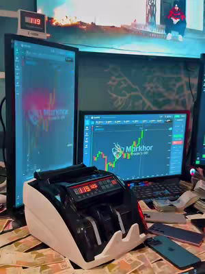 The image shows a desk covered in money, with computer monitors displaying trading charts. A money-counting machine sits in the foreground, with monitors displaying market data and a person (possibly the creator) in the background. The lighting is bright, focusing on assets.
