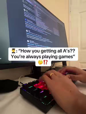 A person is shown typing on a keyboard that has a screen with text behind it, on their right side is a computer monitor with text displayed on it. A text overlay is added to the bottom: ‘: “How you getting all A’s?? You’re always playing games” 😳!?’. The hands are in focus and the keyboard is multicoloured red and blue. The background is out of focus with light-colored walls.
