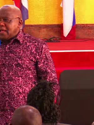 A man in a patterned shirt is speaking, with flags and a red stage in the background. Other people are partially visible in the foreground.