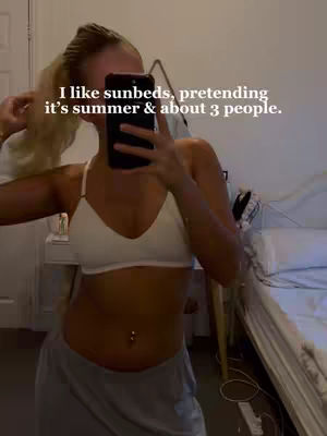 A woman is taking a mirror selfie, her face mostly obscured by the phone she is holding. She has blonde hair pulled back in a high ponytail and is wearing a white bra and gray sweatpants. She has a belly button piercing. The background is a bedroom with a bed visible.