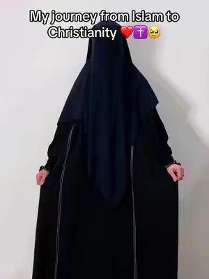 A figure wearing a black niqab and robe stands against a plain light background. The person is seen from the waist up, with their hands held near their waist. Text overlayed on top reads, "My journey from Islam to Christianity," along with heart, cross, and praying hands emojis.