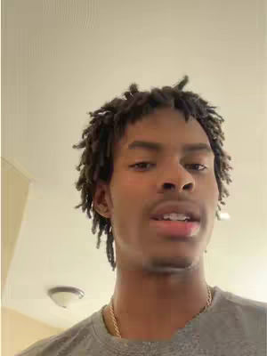 A young Black man with short dreadlocks is looking at the camera while talking. He wears a gray t-shirt and a gold chain.