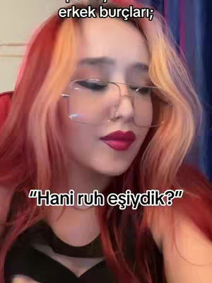 A close-up shot featuring a woman with bright red and auburn hair parted to the side, wearing large-frame glasses and bold red lipstick. In the upper left corner, text overlayed with the phrase "erkek burçları". At the bottom, white text, "Hani ruh eşiydik?'" is displayed,  the background is blurred and indistinct.