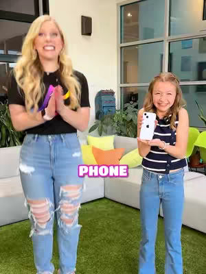 Two females stand in a brightly lit indoor space. The woman on the left is taller with long blonde hair, wearing a black shirt, ripped jeans, and holding her hands together as if clapping or praying. The younger girl on the right is holding a white iPhone and smiling, She wears jeans and a striped top. The background includes a light green wall, a white couch with colorful pillows, and large windows.