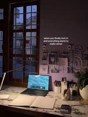 An aesthetic nighttime study setup. A laptop sits open on a desk with a book and other stationary. A window is behind the desk with buildings visible in the background.