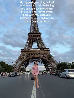 A woman stands confidently on a road in front of the Eiffel Tower, Paris, with cars and pedestrians in the background. She wears a short pink dress and heels. Text overlayed on the image reads: "My biggest green flag is, I will brainwash you into believing you can achieve anything in life, if you spend too much time with me. Because I believe there are no limits to what you can achieve."