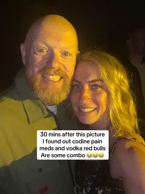 A candid shot of a man and a woman smiling, taken indoors with low lighting. The man on the left has a shaved head, a beard, and is wearing a green shirt. The woman on the right has blonde hair and is wearing a dress or top. Text is overlaid at the bottom.