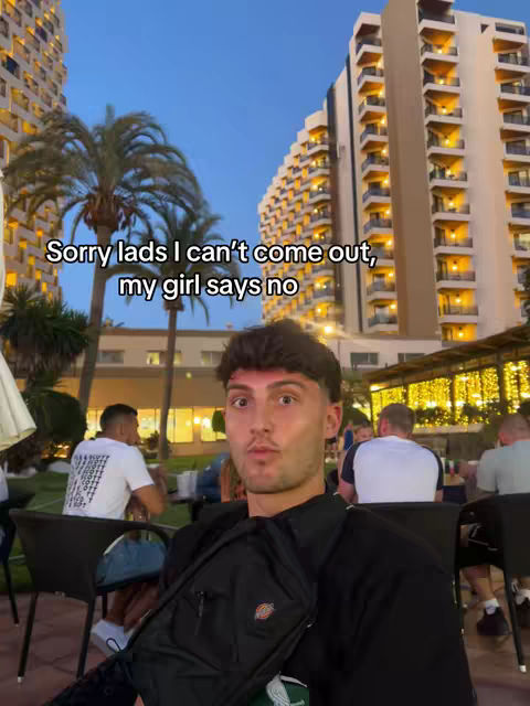 A young man with short brown hair is centered in the frame, looking directly at the camera with a slightly surprised expression. He's wearing a black shirt and has a black bag slung over his shoulder. The background shows a cityscape with tall buildings, palm trees, and people gathered at tables with umbrellas. The sky is a dusky blue.
