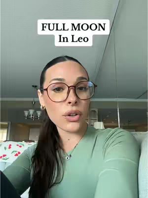 A woman with glasses is centered in the frame. She is looking towards the camera and speaking. Above her head is a white rectangle with the text "FULL MOON In Leo". She's wearing a light green long sleeve top and has her dark hair pulled back. The background is a home environment.