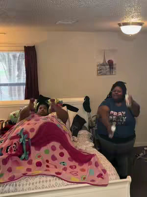 An indoor shot shows two women in a bedroom. One woman is lying in bed under a pink blanket with a cartoon design, while the other woman is partially visible, standing and wearing a t-shirt that reads 'PRAY & SLAY'. The bedroom has a simple layout, with a window, blinds, and a painting hanging on the wall. A chandelier is visible above.