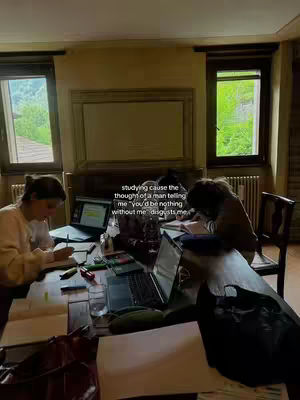 A dimly lit interior shot with two women at a large wooden table. They are studying with laptops and open books. The room has large windows with a view of greenery visible behind them. Text overlay states, "studying cause the thought of a man telling me 'you'd be nothing without me' disgusts me."