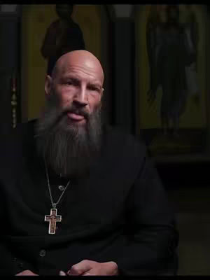 A man with a long gray beard and a black clerical shirt is the central focus. He wears a silver necklace with a cross pendant and his tongue is sticking out slightly. In the background, there are blurred portraits of religious figures. The overall scene is somber and spiritual.