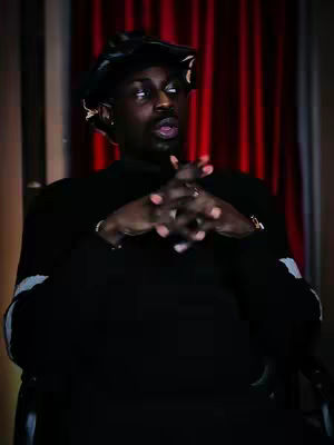 A man in a black sweater and cap is seated with his hands clasped together in front of him. He has a concentrated expression, looking slightly upwards and to the side. The background features a blurred red curtain.