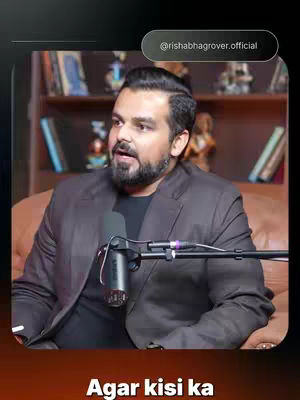 The image features a man in a professional setting, likely a talk show or interview. He is seated, dressed in a dark suit jacket over a black shirt, with a microphone positioned in front of him. His facial expression and hand gestures suggest he is speaking or engaged in conversation. The background shows a bookshelf with books and decorative items, indicating a studio or office environment. Text overlays on the image provide additional context.