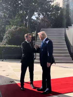 Two men, presumably political figures, are shaking hands on a red carpet. They are standing in front of a building with white walls and a staircase leading up. The setting appears to be outdoors, with trees and greenery visible in the background. One man, possibly representing the United States, is wearing a dark suit with a light-colored tie. The other man is wearing a similar suit but has a different hairstyle. The handshake is the central focus, suggesting a moment of diplomacy or agreement.