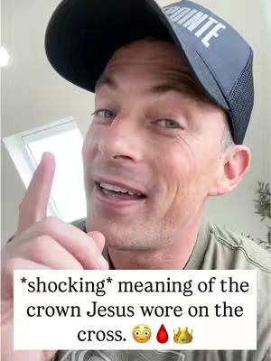 A man with fair skin is shown from the chest up, smiling and pointing his index finger upwards towards the left. He wears a dark blue baseball cap with white text on the front and a gray shirt. The background is blurred and indistinct. Added text at the bottom in a white rectangle includes the words '*shocking* meaning of the crown Jesus wore on the cross', along with three related emojis.