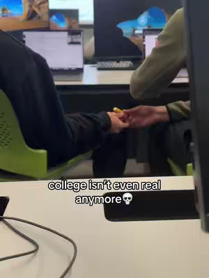 The image shows a scene inside a university or college setting. Two individuals can be seen, one appearing to be seated while the other partially visible. The focus is on the point when the two individuals are holding hands. In the background, computer monitors are visible. A text overlay states, 'college isn't even real anymore' with a skull emoji following it.