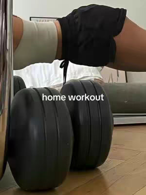 A woman performing a plank exercise, with the upper body visible from the waist up on a neutral-colored top with black shorts. Two black exercise wheels are in the foreground with the text overlayed that says home workout.