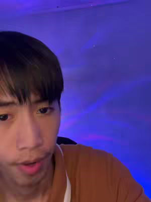 A young man with dark hair and a brown shirt is looking directly at the camera. His expression is open with a slight smile. The background features a purple and blue gradient, possibly from colored lighting, and the focus is on the actor's face.