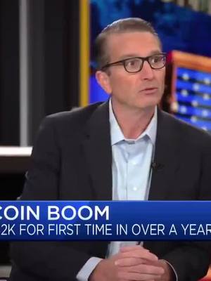 A man in a dark blazer, light blue shirt, and glasses is seated in an interview setting, with a caption reading 'COIN BOOM' at the bottom of the screen.
