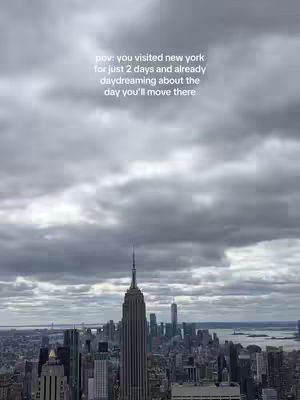 An aerial view of New York City on a cloudy day, showcasing the Empire State Building prominently. The cityscape stretches towards the horizon beneath a sky filled with gray clouds. The buildings are densely packed, and the color palette is subdued with shades of gray and brown.