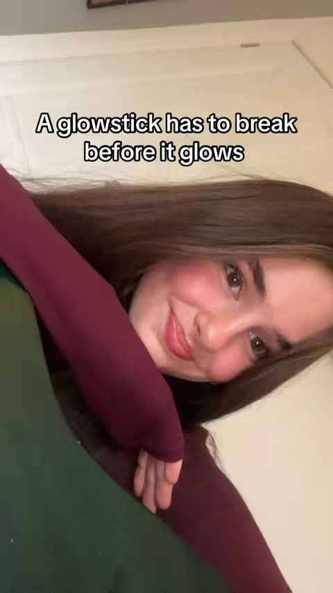 A young woman with long brown hair and rosy cheeks is seen in a close-up shot. She's wearing a burgundy long-sleeved shirt and is slightly off-center with a blurred green background. The text overlay reads: "A glowstick has to break before it glows."