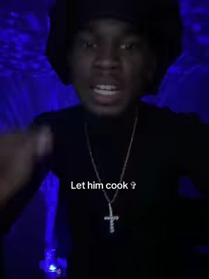 A selfie-style video featuring a young Black person in a dark setting, wearing a black hat, turtleneck, and a cross necklace. Text overlay says 'Let him cook'. The background is a blurred blue. The person is looking at the camera and making a speaking gesture.