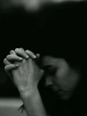 A black and white photo showing a person's hands clasped in prayer, the person's face is partially visible in the shadows.
