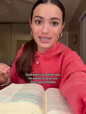 A young woman with long brown hair and light skin is the focus, sitting indoors and looking directly at the camera. She wears a red hoodie and gold hoop earrings, and has braces visible. She’s holding an open book, likely a bible, with its pages open in front of her. Text overlayed on the image says, "God wants to deliver you, he wants to save you from your troubles."