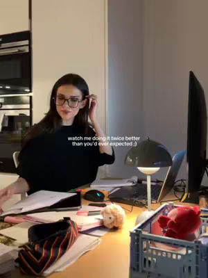 A woman in glasses sits at a desk, studying. The desk is cluttered with papers, a laptop, a computer monitor, and various items. The woman is the central focus, appearing to be engaged in academic work. The setting is illuminated with warm lighting, and a hook is added on the image.