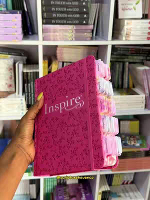 A hand holding a pink book in front of a shelf filled with books. The book has the word "Inspire" in white script on the cover. The background includes white shelves filled with other books.