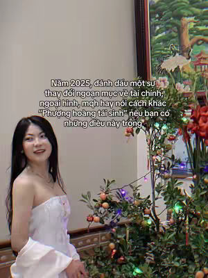 A woman in a white dress stands in front of a background with a picture frame and a plant with fruits and lights and text overlay.