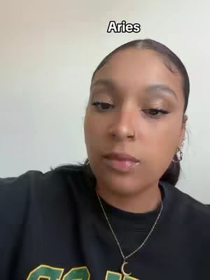 A woman with dark hair and a black shirt is centered in this selfie-style image. The text “Aries” is placed above her head. She has a neutral expression and appears to be looking at the camera. The background is plain and undetailed.