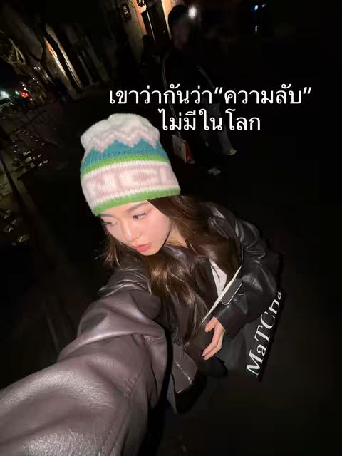 A selfie-style photo taken outdoors at night. The main subject is a young woman wearing a colorful beanie, facing the camera with a neutral expression. The background is dark, with blurred buildings and lights, suggesting an urban setting. The lighting is dim, casting shadows and highlighting the subject’s face and the beanie. Text in Thai overlayed on the image.