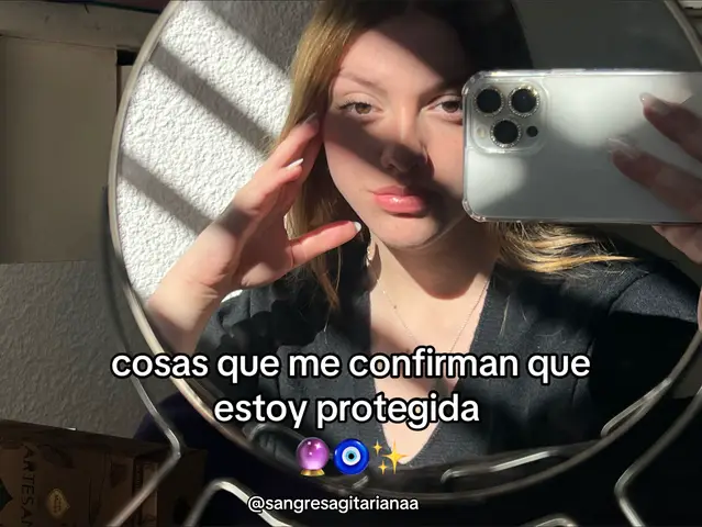 A woman is taking a selfie, holding the phone in front of a mirror. The mirror has a decorative, possibly ornate, frame. The background includes a textured white wall, part of the frame with light and shadow and a glimpse of a different setting. The woman has light hair and is wearing a dark top. Overlay text indicates themes of personal protection and spirituality, with additional symbols such as a purple crystal ball, an eye, and stars.
