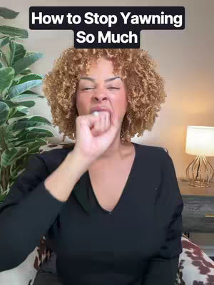 A person with curly hair and a black top yawns, covering their mouth with a hand. The background includes part of a plant, a wall, and a lamp.