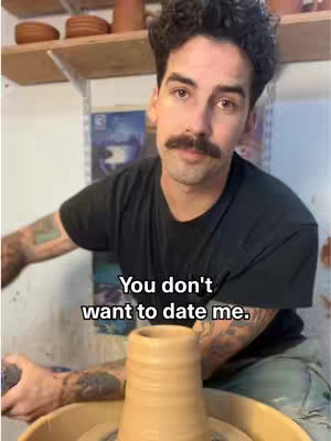 A man in a black t-shirt with tattoos on his arms is seated in front of a pottery wheel. The background includes shelves with pottery and a whiteboard. Text overlayed across the bottom reads, 'You don't want to date me.'