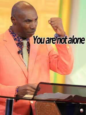 A man in a peach suit is standing behind a lectern, with the text "You are not alone" overlaid on the image. He has a serious expression and his right fist is clenched.