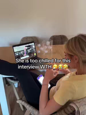 A woman is seated at a table with her legs extended, holding a phone and appearing relaxed. Her laptop displays a video call with what appears to be a person in a suit. The room has a neutral color palette with a wooden table, a cream-colored wall, and windows. The woman is wearing a yellow t-shirt.
