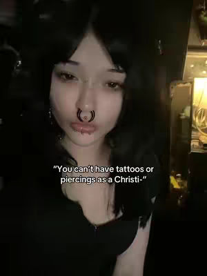 A close-up shot features a person with pale skin, dark hair, and multiple facial piercings (a septum ring and lip piercings). The person's eye expression is neutral, and the background is dark. Overlayed text states, "You can't have tattoos or piercings as a Christi-".
