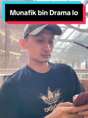 A man wearing a baseball cap and a black shirt with a white logo is looking down at a phone he's holding, the phone is in his right hand. Behind him is a window with daylight visible. Overlay text at the top.